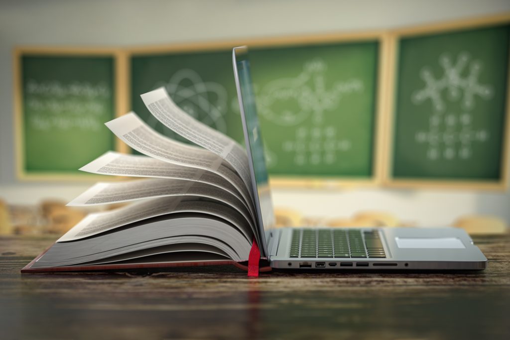 Otwarty laptop stojący na drewnianym stole, którego ekran płynnie przechodzi w kartki papierowej książki. W tle widać rozmazany obraz tablic szkolnych z narysowanymi wzorami chemicznymi i matematycznymi, co symbolizuje połączenie edukacji tradycyjnej z cyfrową.
