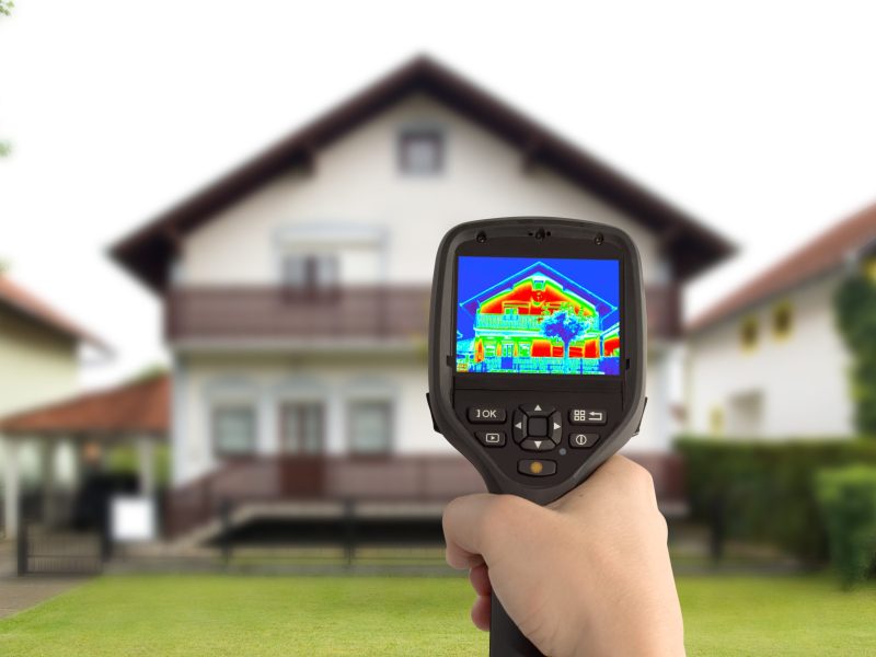 Widok na ekran kamery termowizyjnej trzymanej w dłoni, która pokazuje obraz piętrowego domu jednorodzinnego. Kolory na wyświetlaczu wskazują miejsca ucieczki ciepła z budynku, podczas gdy w tle widać rzeczywisty, lekko rozmazany widok tego samego domu.
