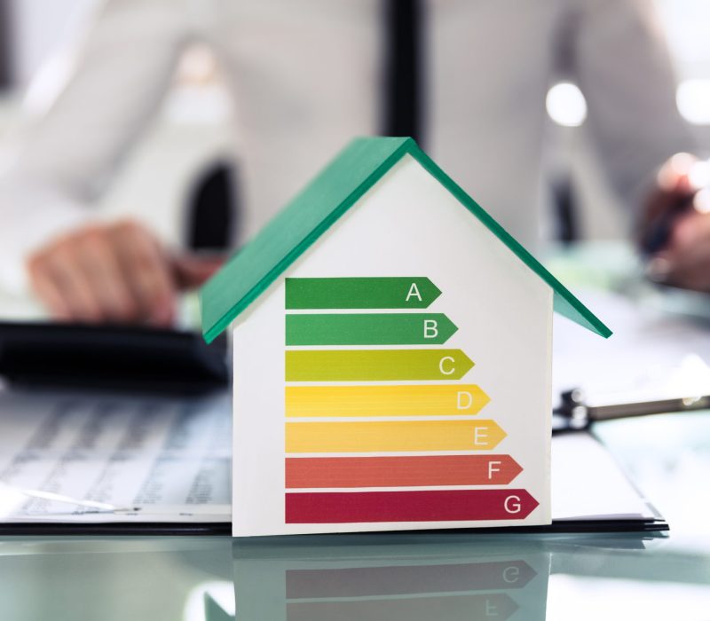 Model białego domku z zielonym dachem, na którego frontowej ścianie umieszczono kolorową etykietę klas energetycznych od A do G. W tle widać niewyraźną postać osoby w koszuli pracującej przy biurku z dokumentami i kalkulatorem.